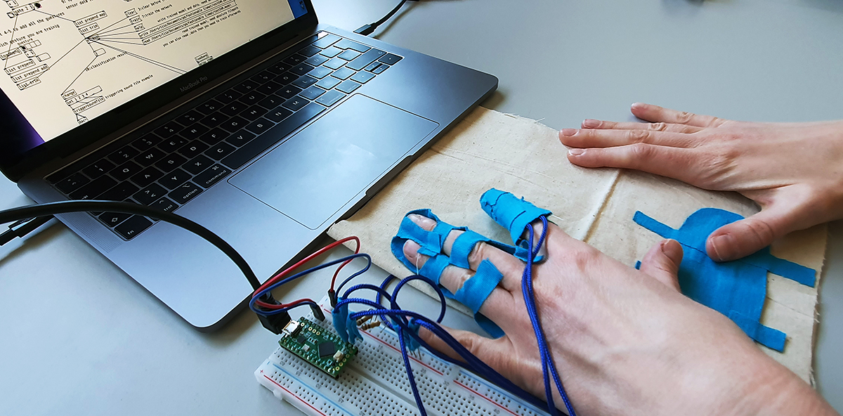 A laptop with some visual coding, microcontroller and hands with blue tape around them.