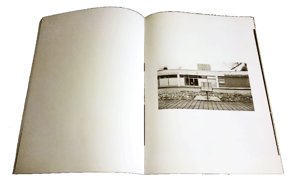 Open zine, one black&white photo of
	urban area with empty chair for one person, otherwise the spread is empty.